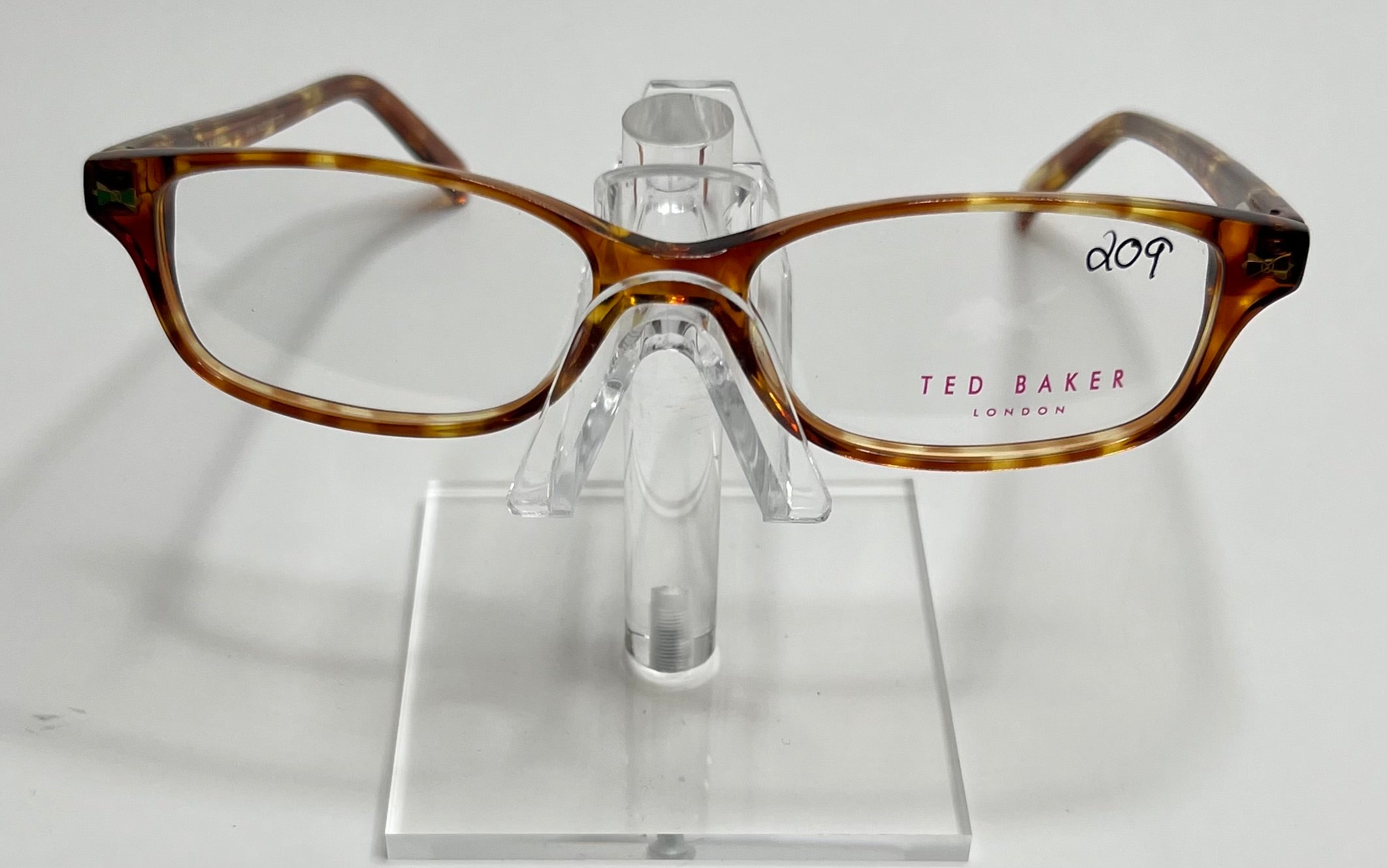 A pair of Ted Baker B705 ladies plastic eyeglass frames in a golden tortoise shade, displayed on a stand.