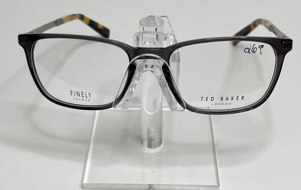 A pair of Ted Baker TFM004 men's eyeglasses with a sleek gray front and tortoise temples displayed on a stand.