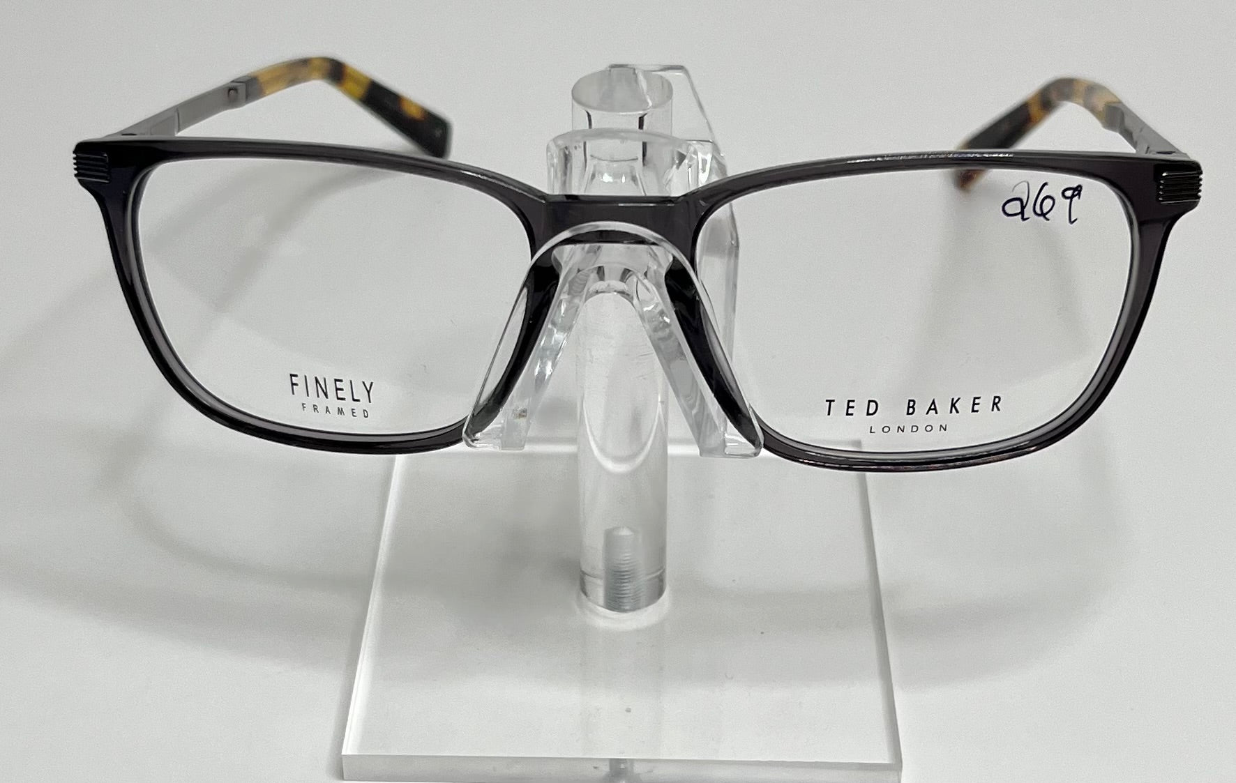 A pair of Ted Baker TFM004 men's eyeglasses with a sleek gray front and tortoise temples displayed on a stand.