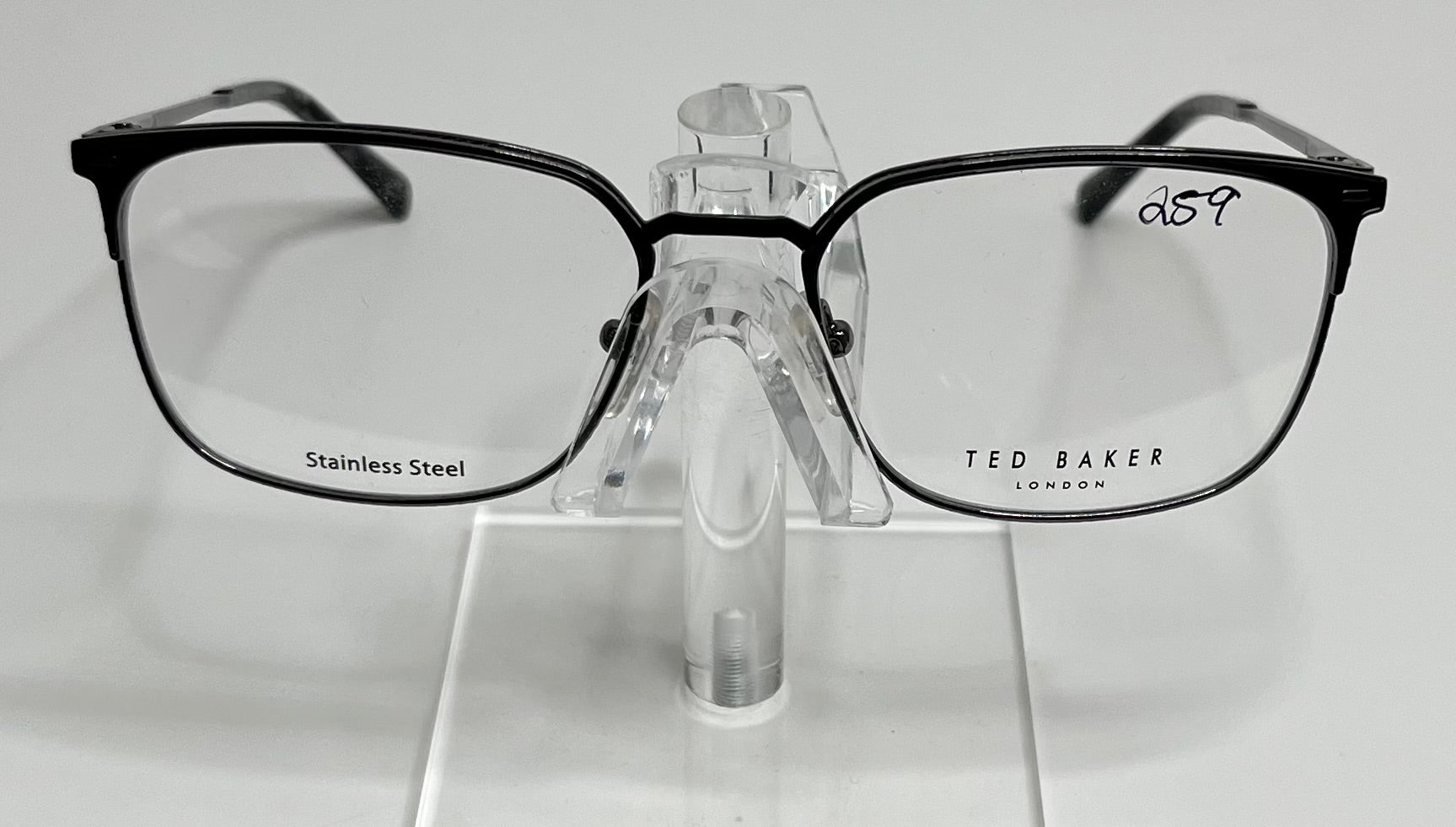 A sleek gunmetal stainless steel eyeglass frame by Ted Baker displayed on a stand.