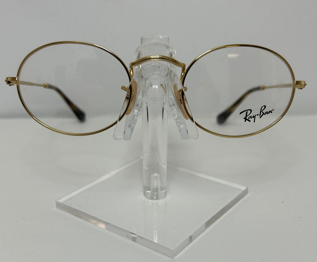 Oval round eyeglasses with a gold metal frame and clear lenses, displayed on a transparent acrylic stand. The Ray-Ban logo is printed on the lower right lens.