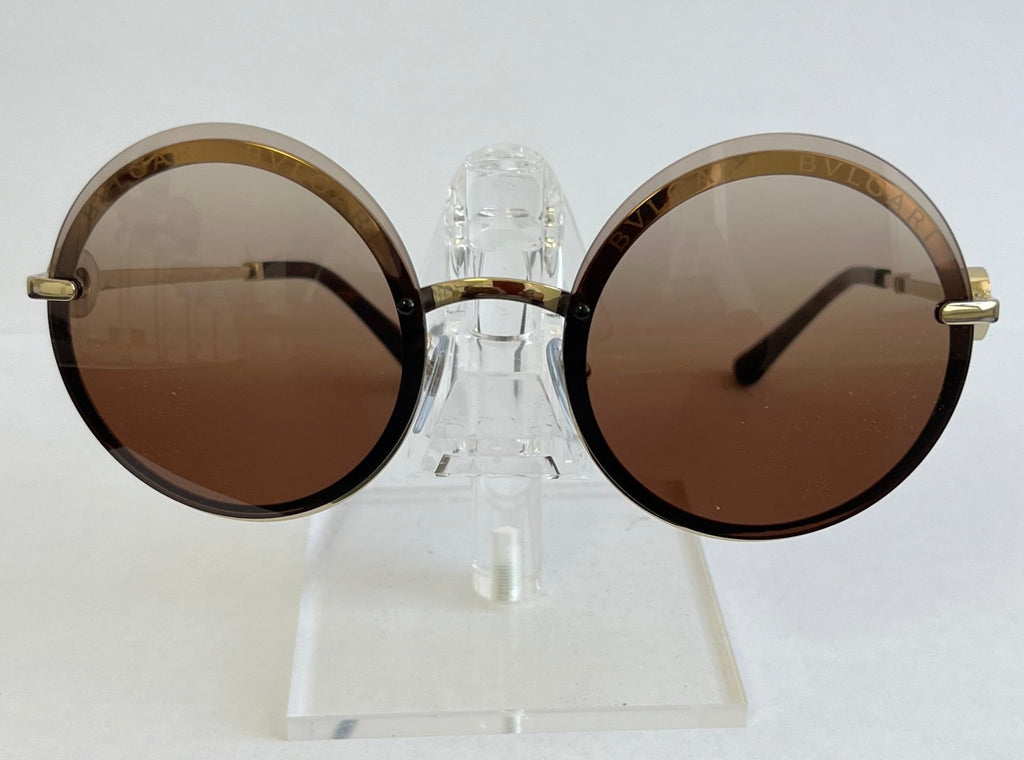 Round sunglasses with gold frames and brown lenses on a clear stand against a white background